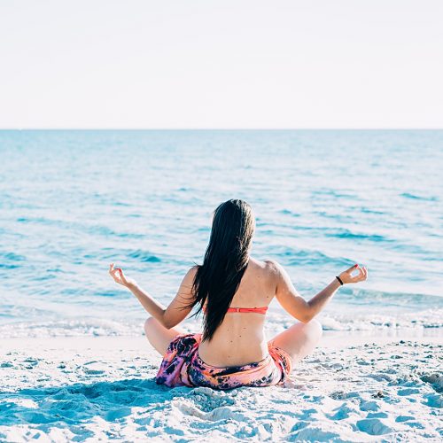 Back view of young beautiful woman doing yoga at the beach in summertime in lotus position- relax, meditation, spiritually concept